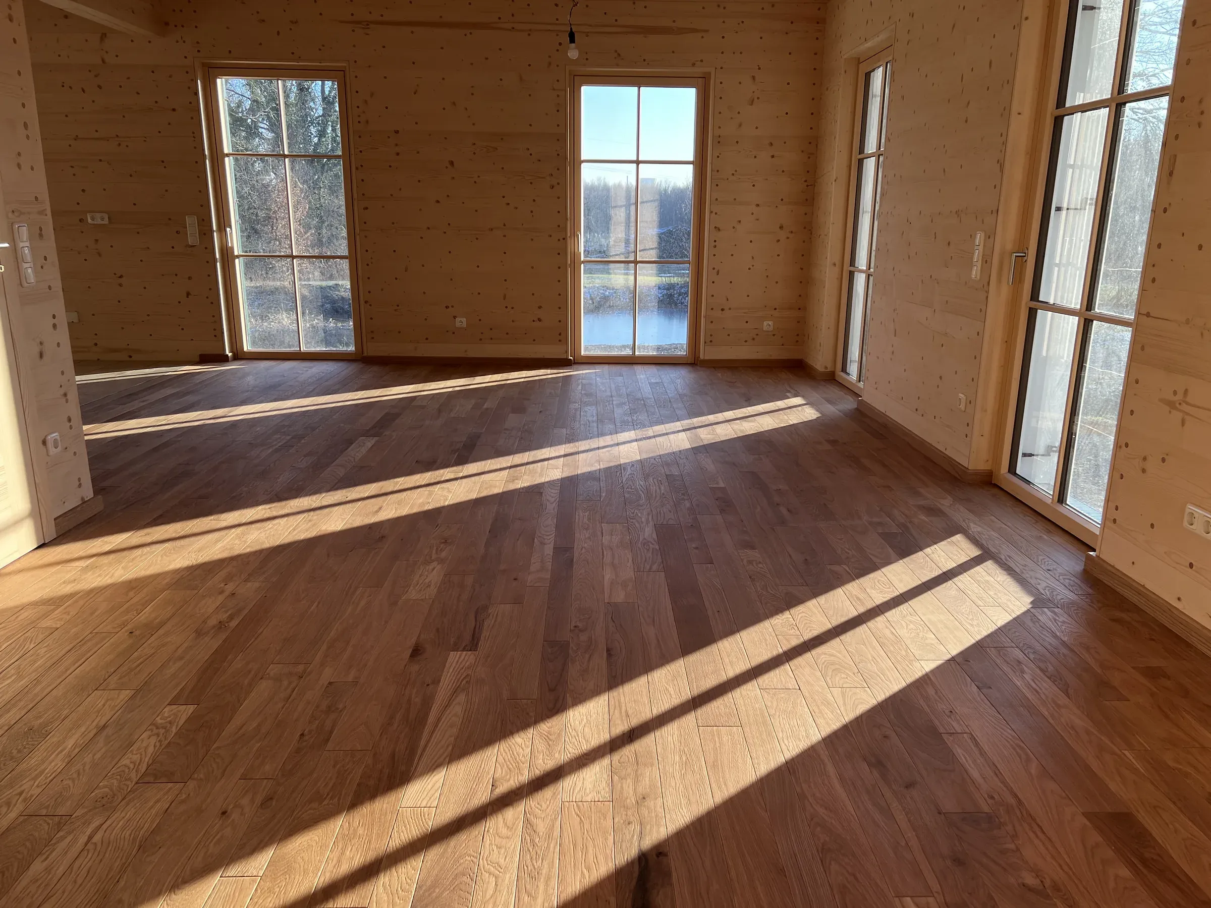 Wohnraum mit holzverkleideten Wänden, bodentiefen Holzfenstertüren mit Sprossen und Dielenboden im Streiflicht