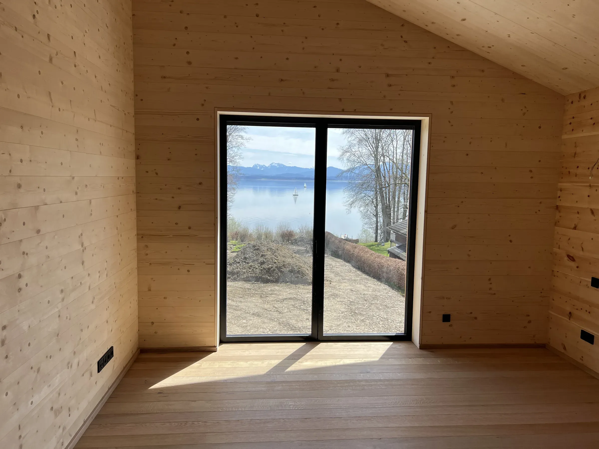 Holzverkleideter Raum unter einer Dachschräge mit großer, dunkel gerahmter Fenstertür und Blick auf einen See und Berge