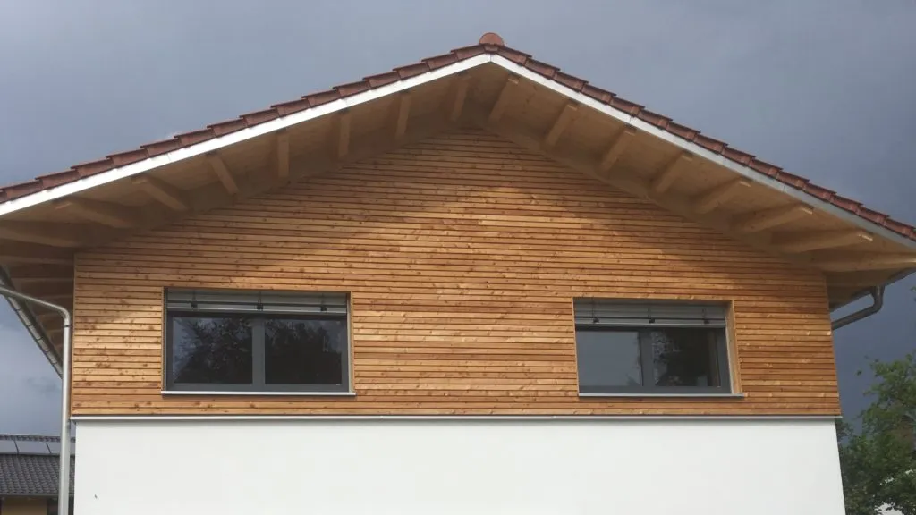 Giebelfassade mit horizontal verlegten Holzlamellen unter einem Ziegeldach, zwei Fenster mit Rollläden