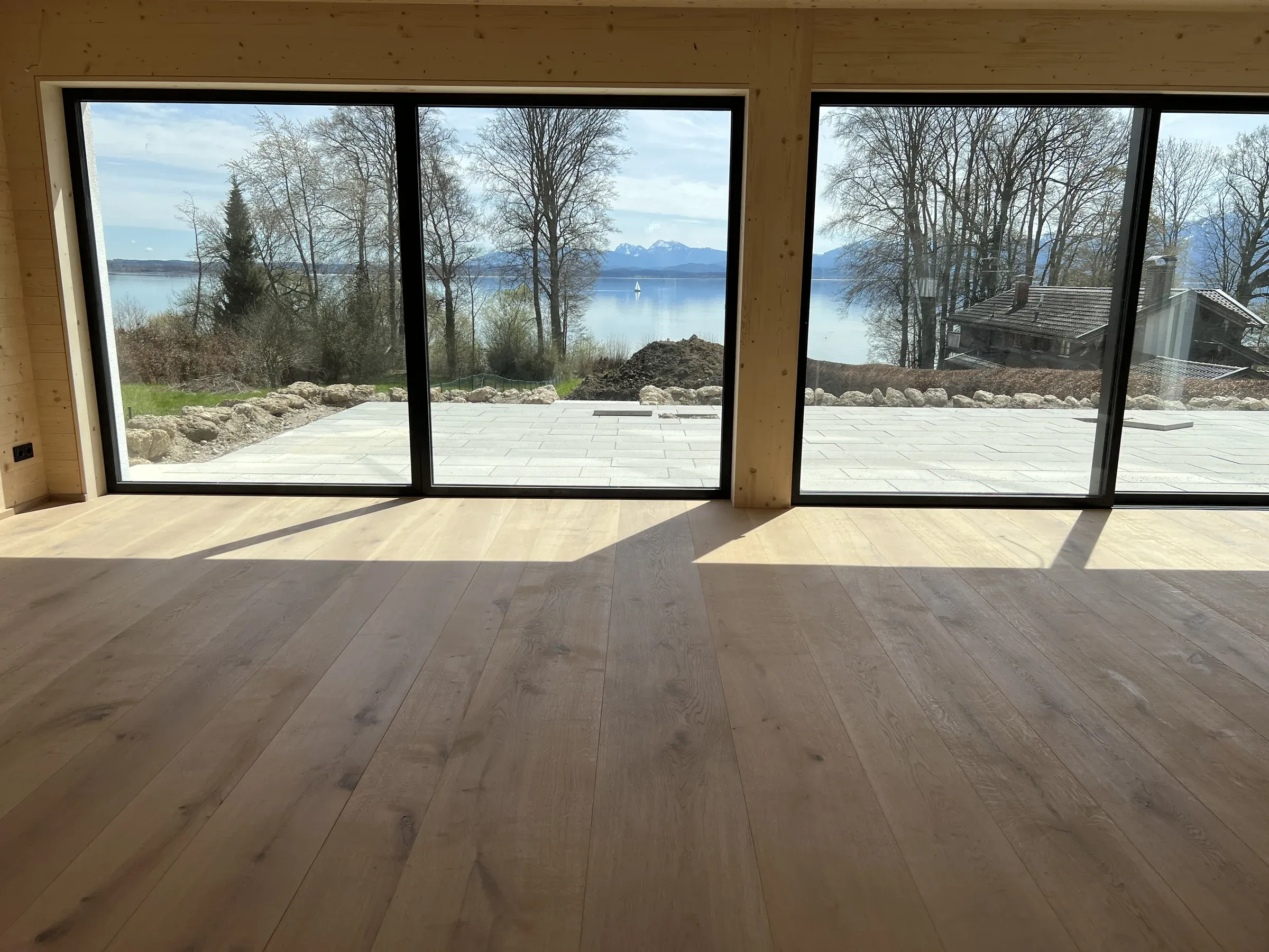 Wohnraum mit breiten hellen Landhausdielen, Holzwänden und großen dunkel gerahmten Fenstertüren mit Blick auf See und Berge